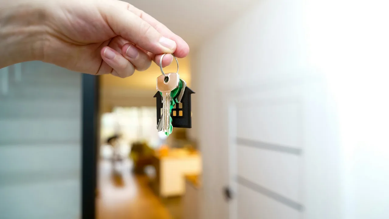 A hand holding house keys with a house-shaped key ring in a new home hallway